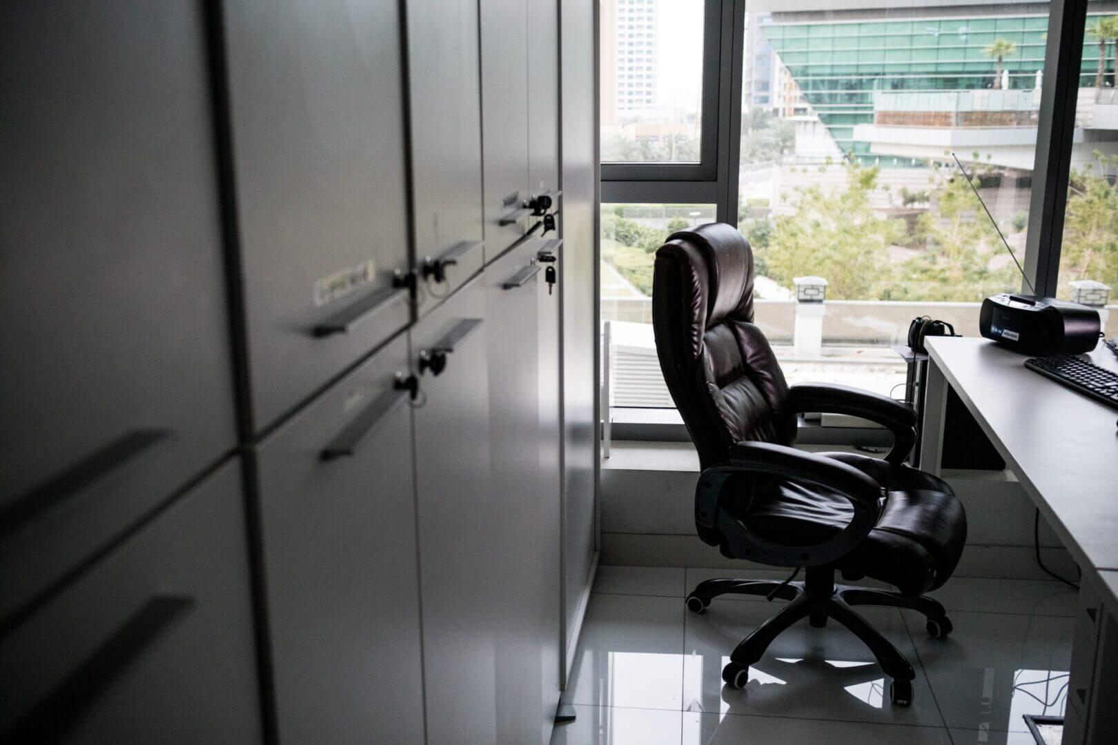 Office space with empty leather chair, desk, and cabinets near large window; modern business environment.