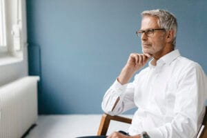 Senior manager in office chair gazing out window, deep in thought, wearing glasses and white shirt.